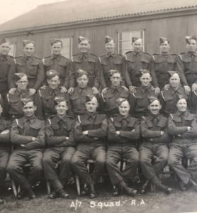 The photo shows a group of men from the Royal Artillery lined up in three rows; the front row is seated while the middle row is stood, and the back row is stood raised above the middle. Their Royal Artillery cap badges are visible. The caption reads: 'A/7 Squadron, Royal Artillery'. 