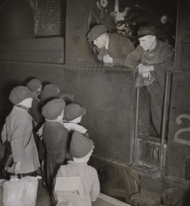A group of six evacuees stand on the platform of a London station in March 1941, speaking to the engine driver. The evacuee nearest the camera is wearing their gas mask box on their back, like a backpack. © Hulton Archive/Getty Images