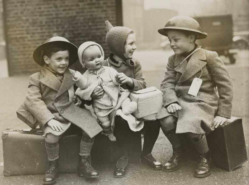 An undated image of three evacuee children during WWII. All three sit on their suitcase, with a girl in the middle holding a toy doll and two small boys on either side, wearing tin helmets. They are carrying gas masks, and their identity tags, affixed to their coats, are visible. One of the tags contains the letters 'L.C.C.', meaning London County Council. Clues like these are useful when researching evacuees and their experiences during WWII. Cars can be seen in the background; the children may be sitting in a school playground or on school property ahead of their train journey to safer parts of the county, known as reception areas. © Hulton Archive/Getty Images