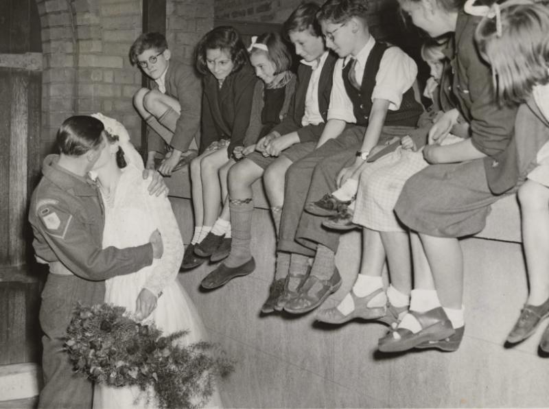 Fred Simpson kisses his bride, Alice Mico, following their marriage on 10 October 1953. Fred was a prisoner of war during WWII and the Korean War. In the image, Fred is wearing his British Army uniform. The insignia on his right arm indicates that he was a lance corporal in the Gloucestershire Regiment. A group of children sat on a wall, look on, smiling at the couple. © Hulton Archive/Getty Images