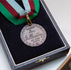 The Dickin Medal. The image shows a commemorative box containing the bronze medal. The medal reads 'PDSA. For Gallantry. We also Serve'. It has a ribbon of three colours: green, dark red and light blue. © PDSA