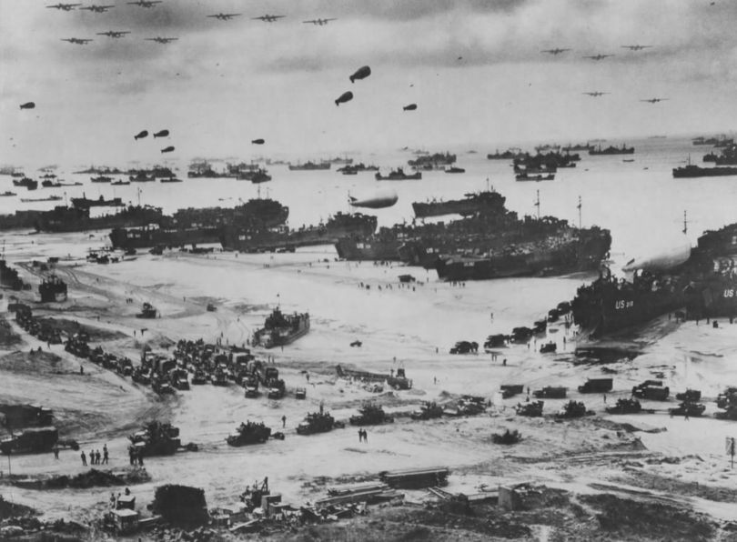 A striking panorama of the Normandy landings on one of the invasion beaches. Aircraft can be seen in the sky with barrage balloons floating out at sea. Landing craft are docked at the shore and troops and vehicles can be seen on the beach. ©NARA