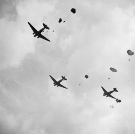 Paratroopers and equipment drop from Dakota Aircraft over the outskirts of Arnhem on 17 September 1944, during the Battle of Arnhem. The image is shot from below and shows three Dakota aircraft against the sky. Paratroopers can be seen in the process of exiting the planes or plunging through the sky with their parachutes open or opening. Small oblong equipment is also visible, descending into the sky and attached to a parachute. © IWM (BU 1162)